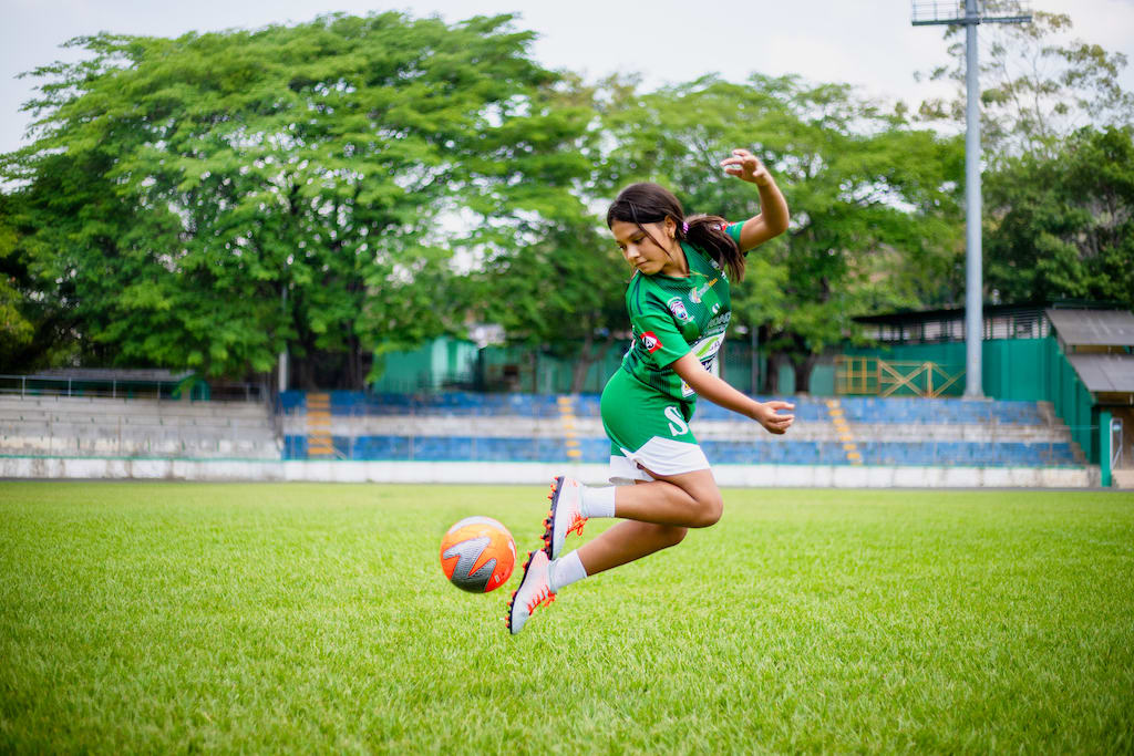Girl in a green soccer jersey kicks a soccer ball behind her