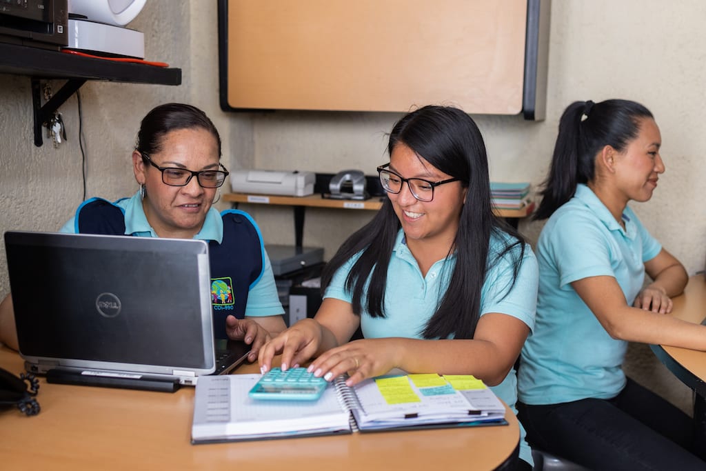 A Compassion centre director and two Compassion students, all wearing light blue uniforms, sit at desks using computers and textbooks to study.