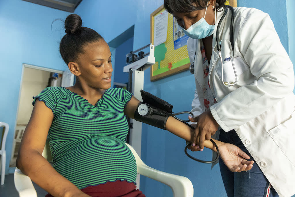 Noelia, in a green shirt, is sitting in a chair while a Survival doctor in a white coat and face mask checks her blood pressure during a routine checkup. Noelia is 38 weeks pregnant.