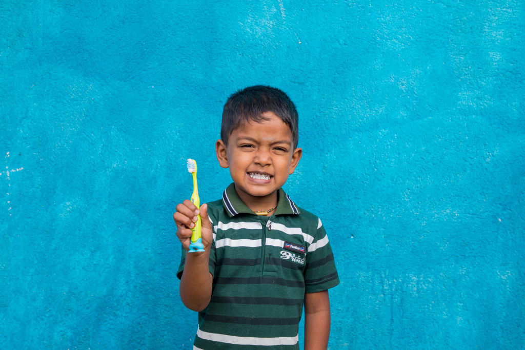 Rakshan wears a green striped shirt and holds up a bright green toothbrush. He smiles with his teeth showing.