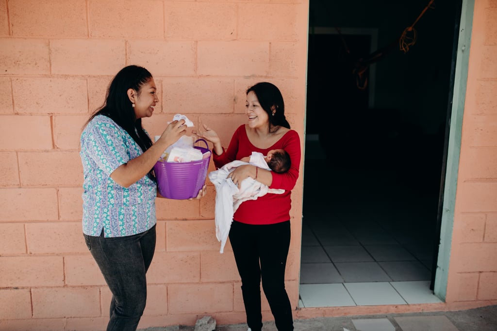 Brenda holds Samantha outside of their home while receiving a purple gift basket of baby goods to keep her healthy from a Compassion staff member.