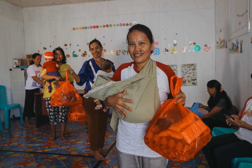 Jeni, Serli and other mothers with their babies are standing in a room at the Compassion centre holding bags of supplies provided by the Survival program.