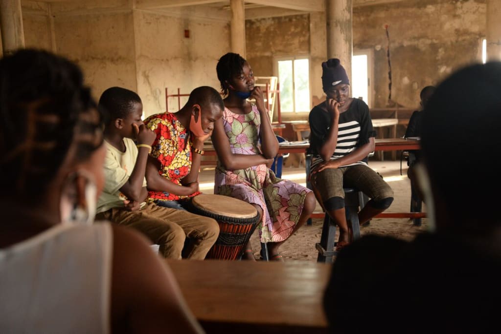 A group of youth who are part of the Cultural Club are participating in story telling. They are all sitting on stools while other children are sitting at tables and watching.
