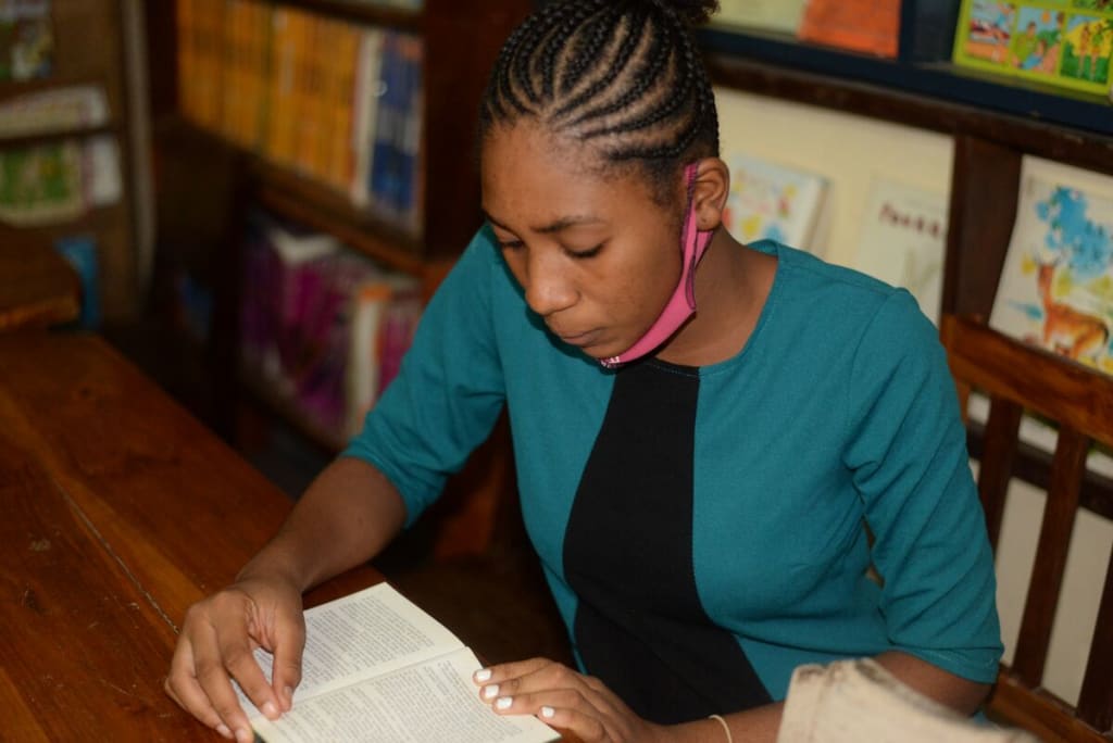Abigael is wearing a black dress with a green jacket, as well as a pink face mask that is down around her chin. She is sitting at a table and is reading a book in the Compassion center's library. Behind her are book shelves.