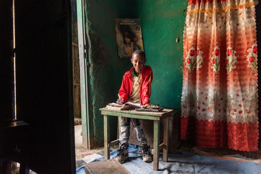 Abel, in a red jacket, is at home sitting at a small green wood desk. The walls behind him are green and there is an orange and red curtain hanging next to him.