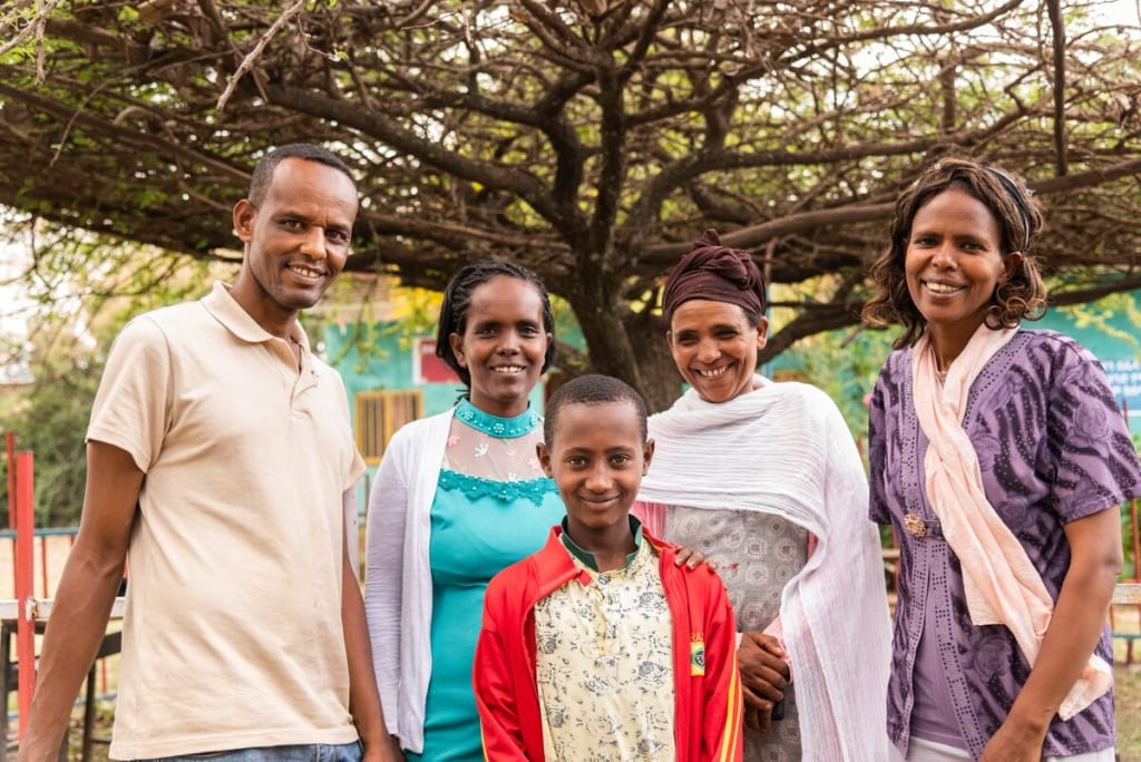 From left to right: Abaya (Project Director), Shahitu (health worker, in a blue dress), Astede (Abel’s mom, in white), Meseret (social worker, in purple) and Abel (in red) are standing outside posing for a picture in front of a tree.