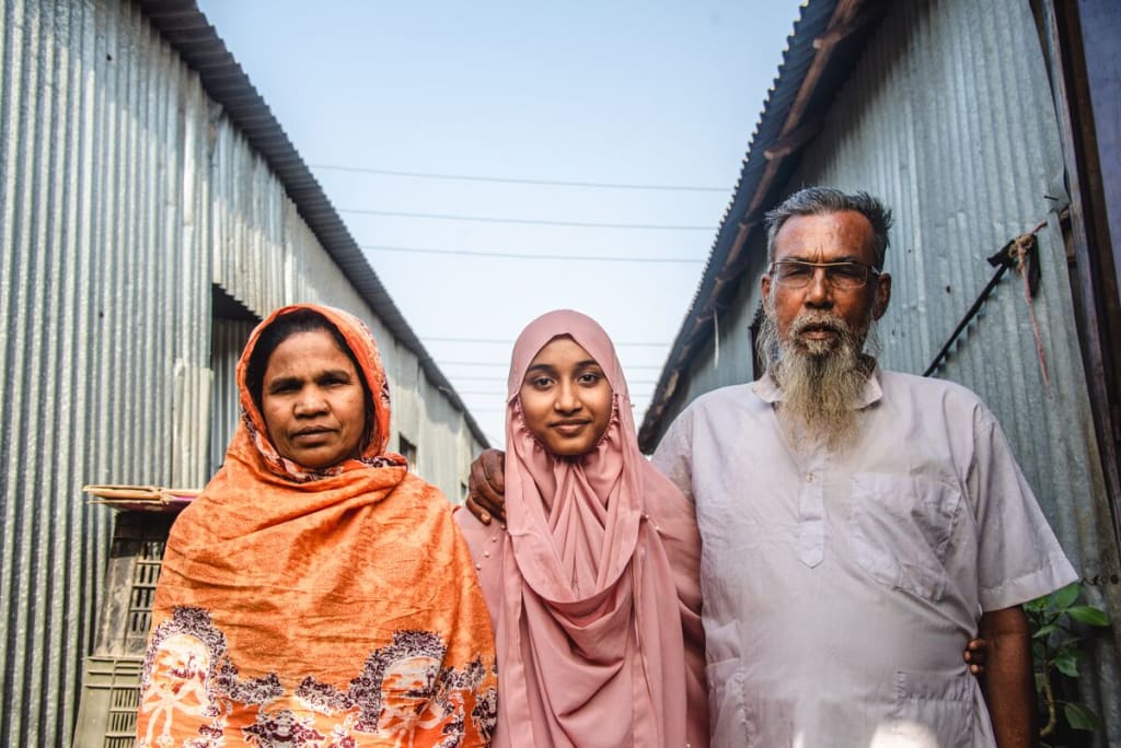 Akhi is wearing a pink dress and head covering. She is standing outside her home with her mother, wearing an orange dress and head covering, and her father, wearing a light gray shirt. They have their arms around each other.