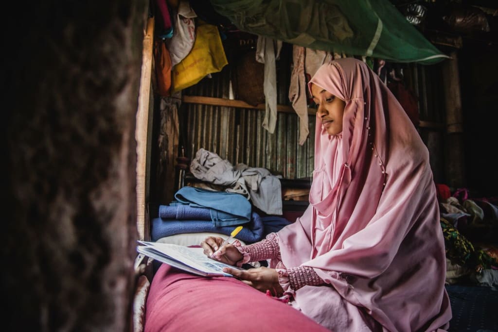 Akhi is wearing a pink dress and head covering. She is sitting in front of a window in her home and is writing a letter to her sponsor.
