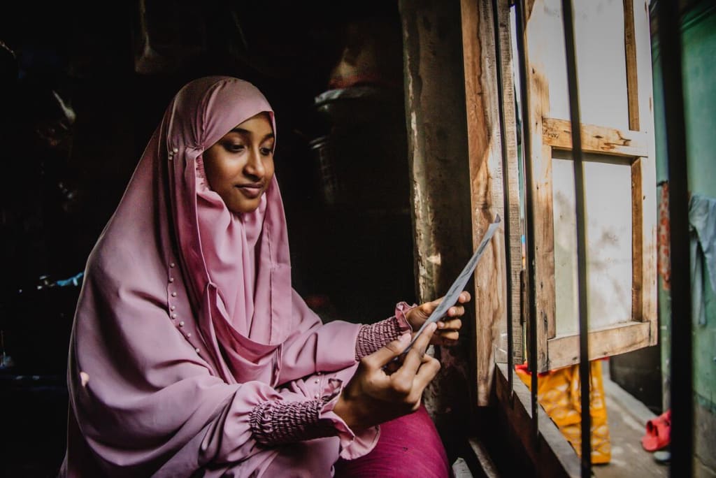 Akhi is wearing a pink dress and head covering. She is sitting in front of a window in her home and is looking at a letter from her sponsor.