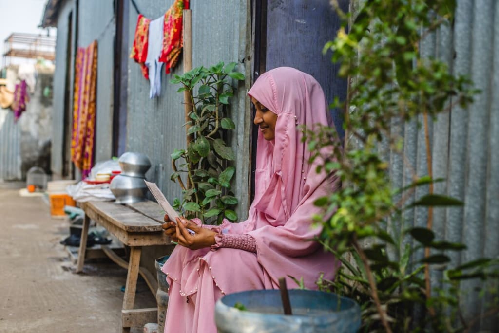 Akhi is wearing a pink dress and head covering. She is sitting in the doorway of her home and is holding a letter from her sponsor. Her home is made of corrugated metal.