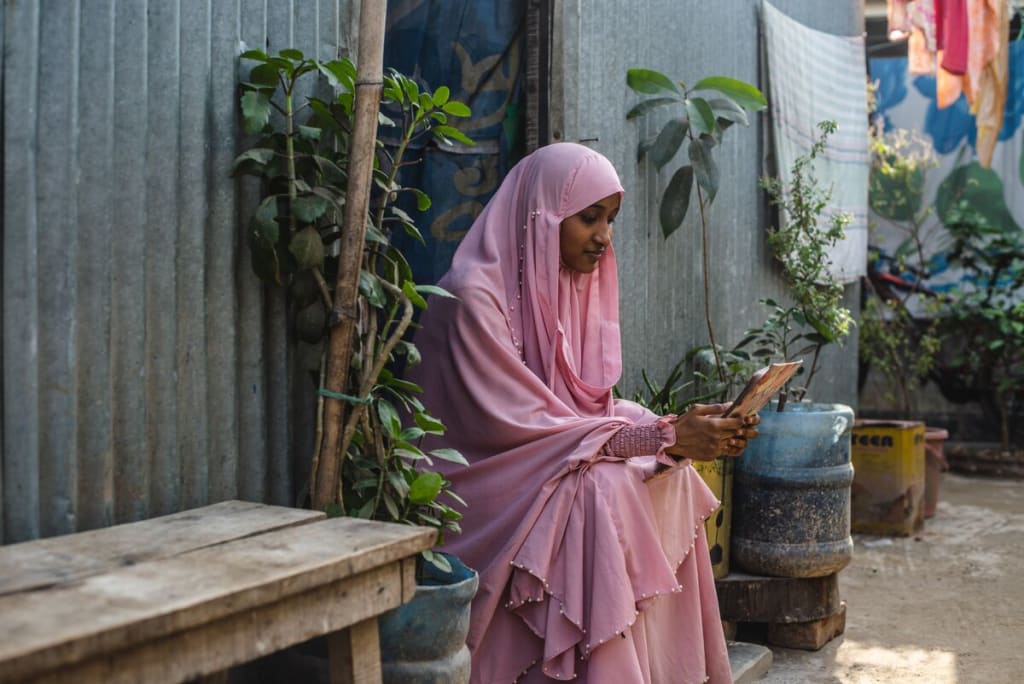Akhi is wearing a pink dress and head covering. She is sitting in the doorway of her home and is reading a letter from her sponsor. Her home is made of corrugated metal.