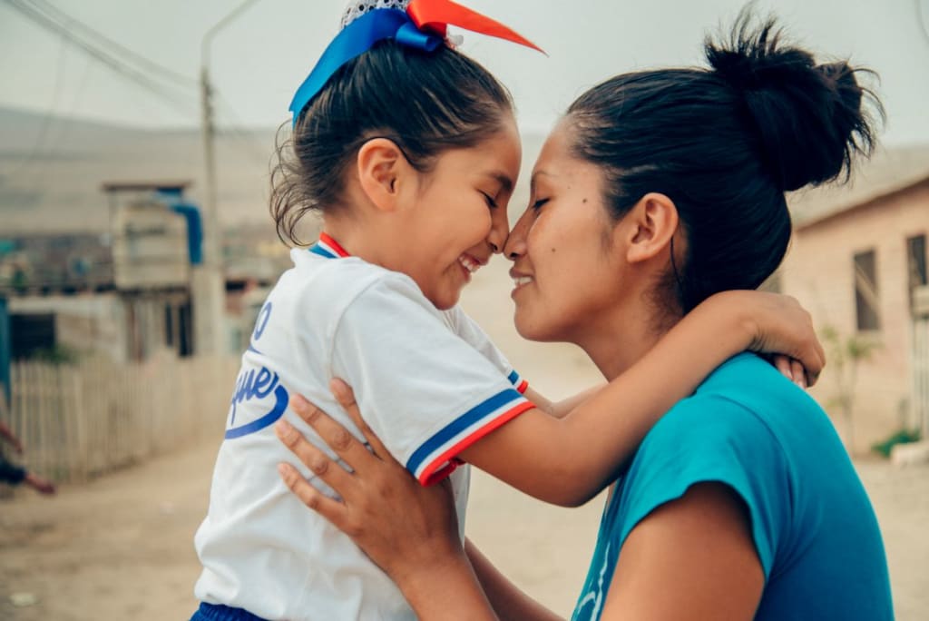 A young girl held by her mom. She is wearing red, white and blue ribbons bows in her hair. She leans in to touch her nose to her mom's nose.