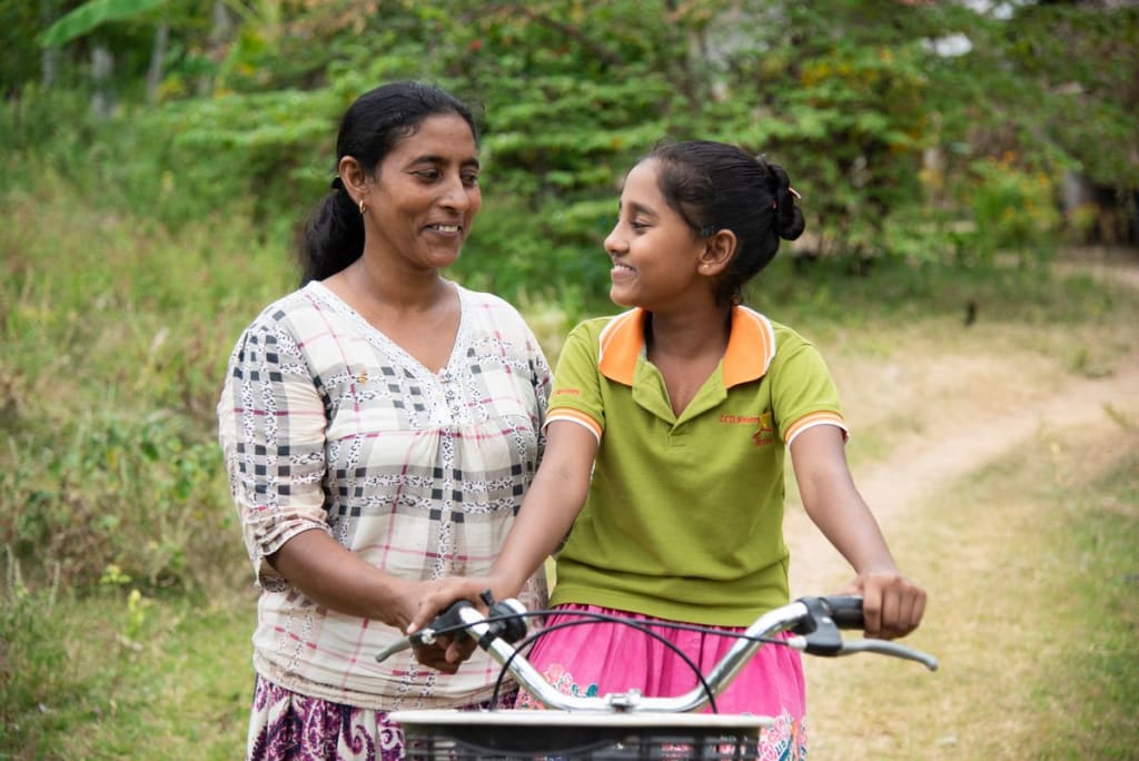 A girl on a bike is wearing a green shirt and a pink skirt. Her mother is wearing a colorful skirt and a white patterned top. They are smiling at each other.