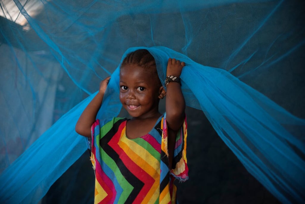 Suzanna, Grace's daughter, is wearing a colorful dress and is sitting under a blue mosquito net. She is smiling at the camera.