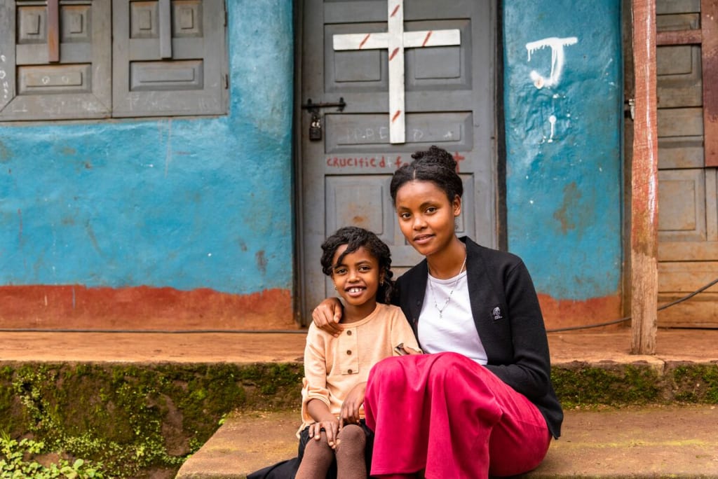 Young woman wearing a pink skirt sits with a young girl on the steps of a church with blue walls