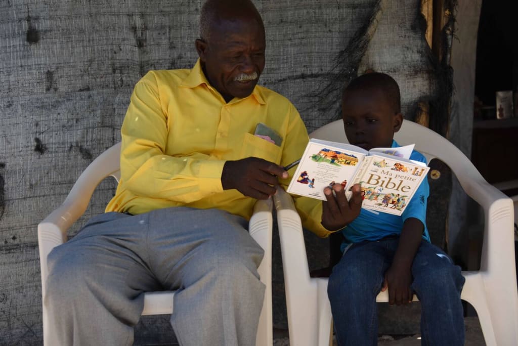A man in a yellow shirt reads to a child in blue on lawn chairs