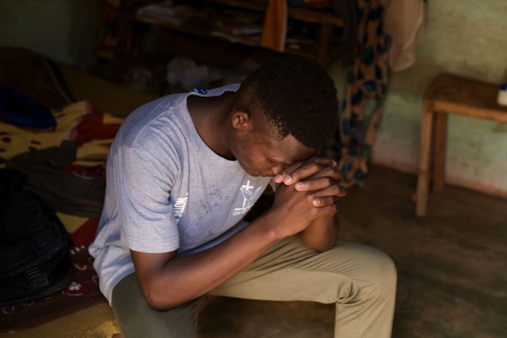 A teenager in a grey shirt prays on a bench