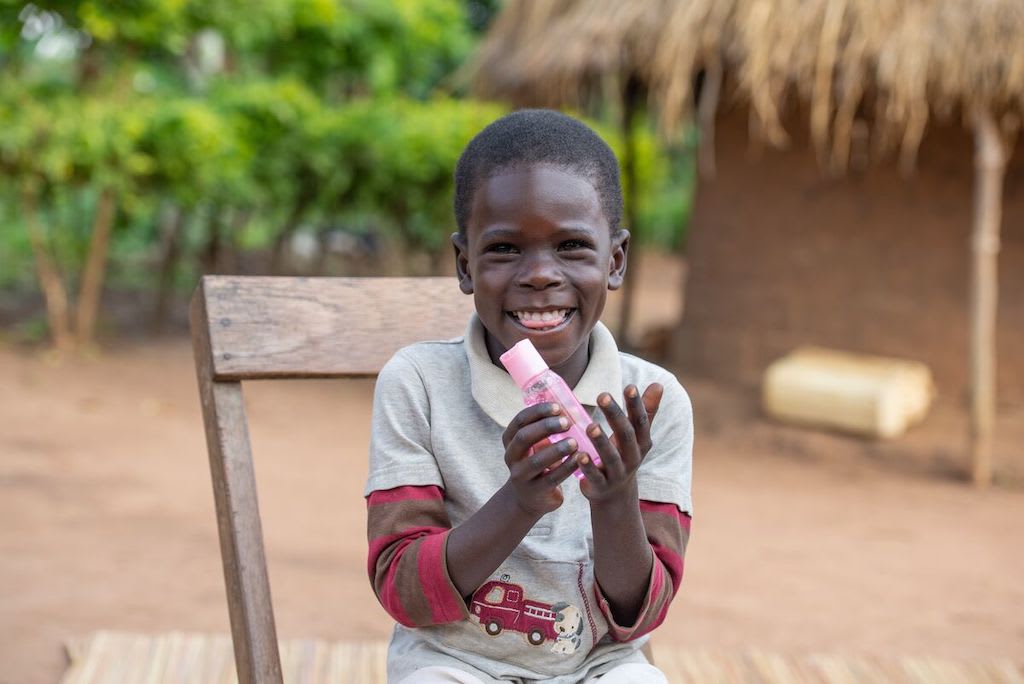 A boy sits on a chair, smiling and holding a pink bottle of hand sanitizer.