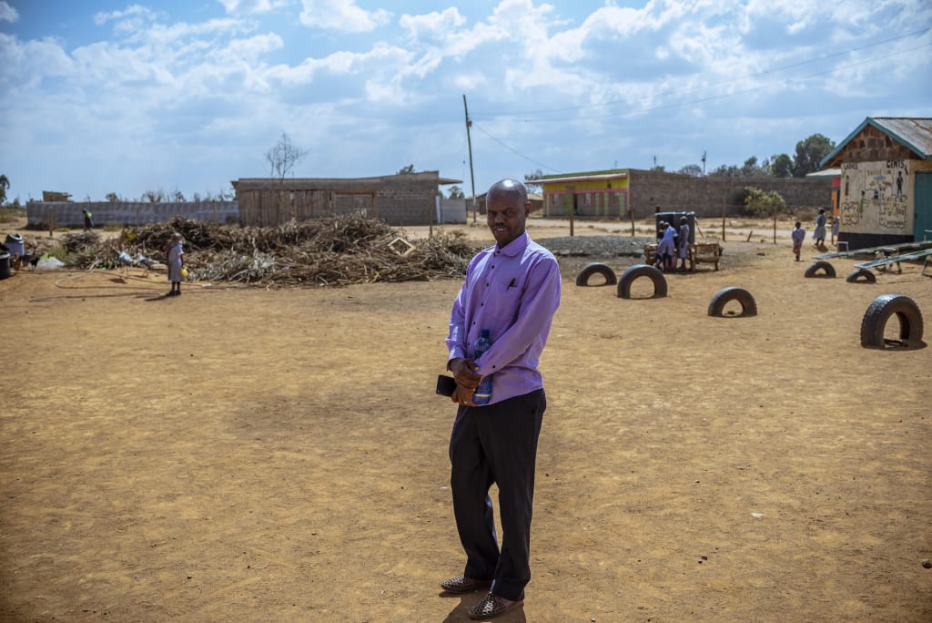 Pastor Richard stands in an open field in his community, wearing dark pants and a light purple shirt.