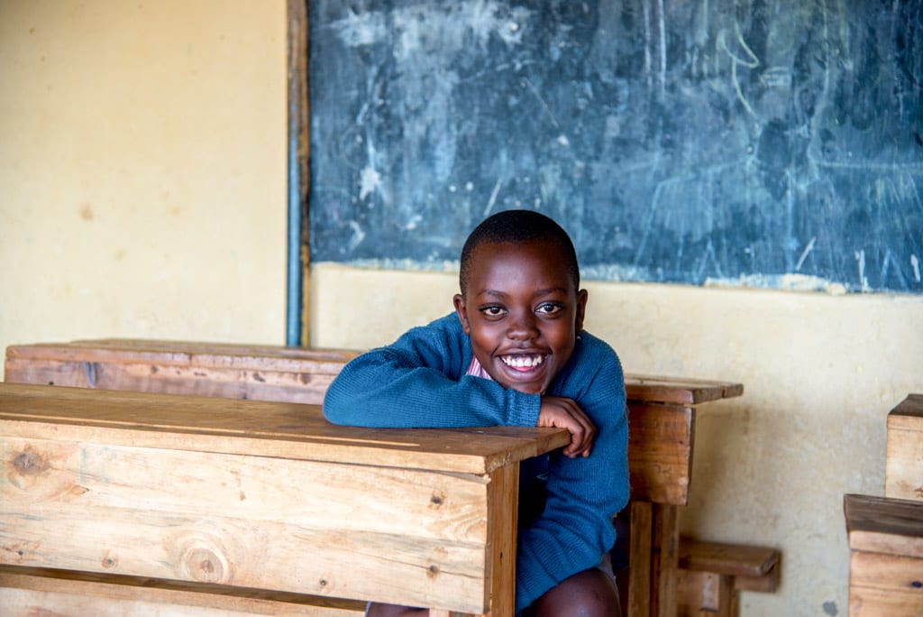 A boy sits a wooden school desk in a classroom and smiles at the camera.