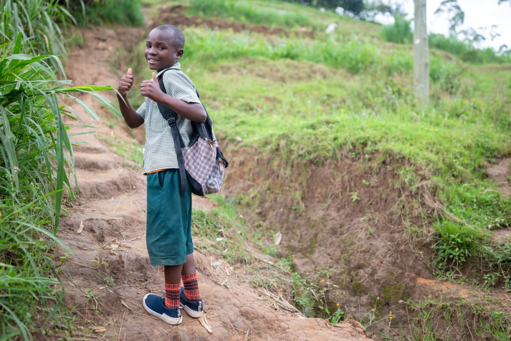 A boy in a tshirt and green pants wears a backpack and gives two thumbs up while standing on a path.