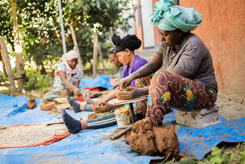A dark skinned woman in grey and brown clothing sits at a pottery wheel, shaping clay which will become useful pottery.