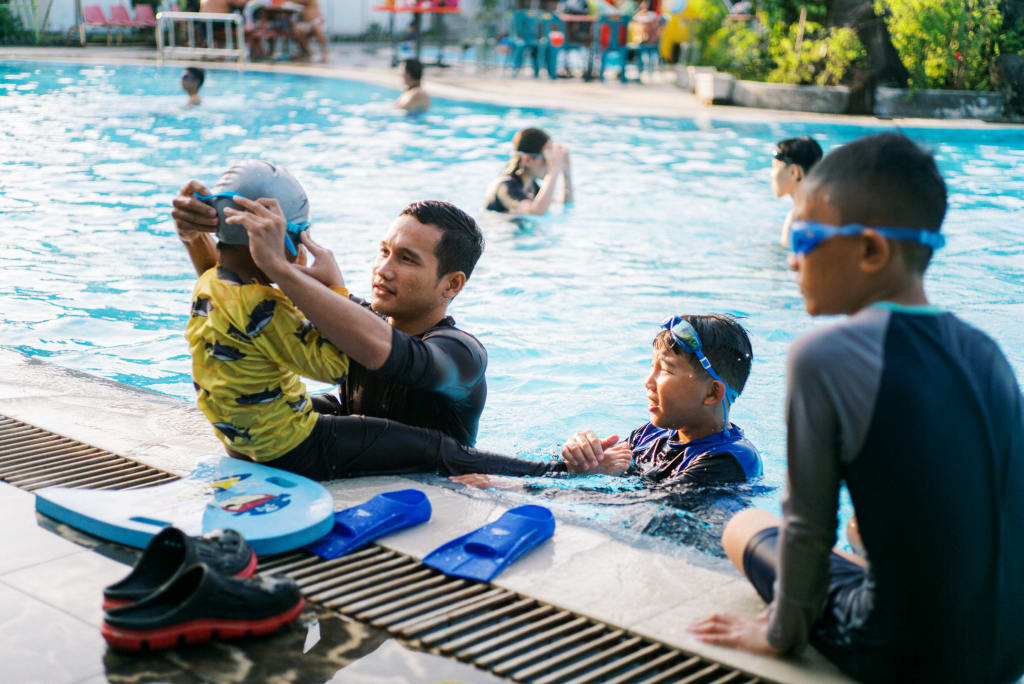 Johanes is at the edge of a swimming pool adjusting a child’s goggles. He is surrounded by his students.