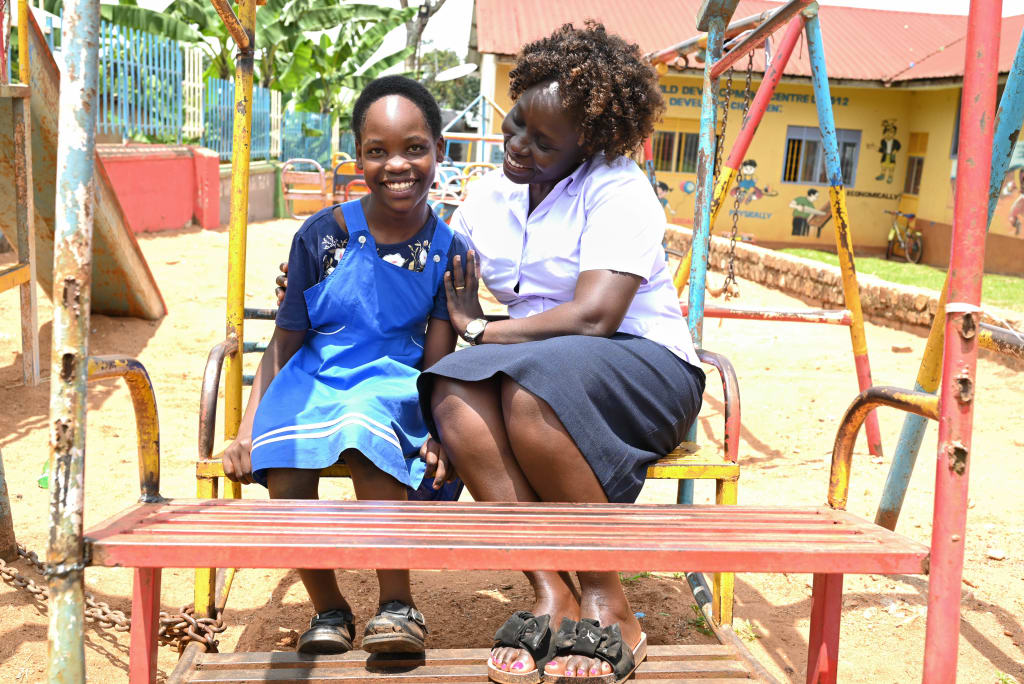 Juliet is outside on the playground at the centre, sitting on a piece of playground equipment with Christine, one of the staff at the church who encourages Juliet in facing her disability.