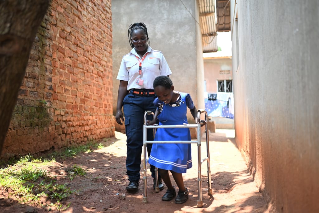 Juliet is outside walking with her mother, Agnes, who is wearing her security guard uniform. Juliet is using a walker to help her overcome her disability.