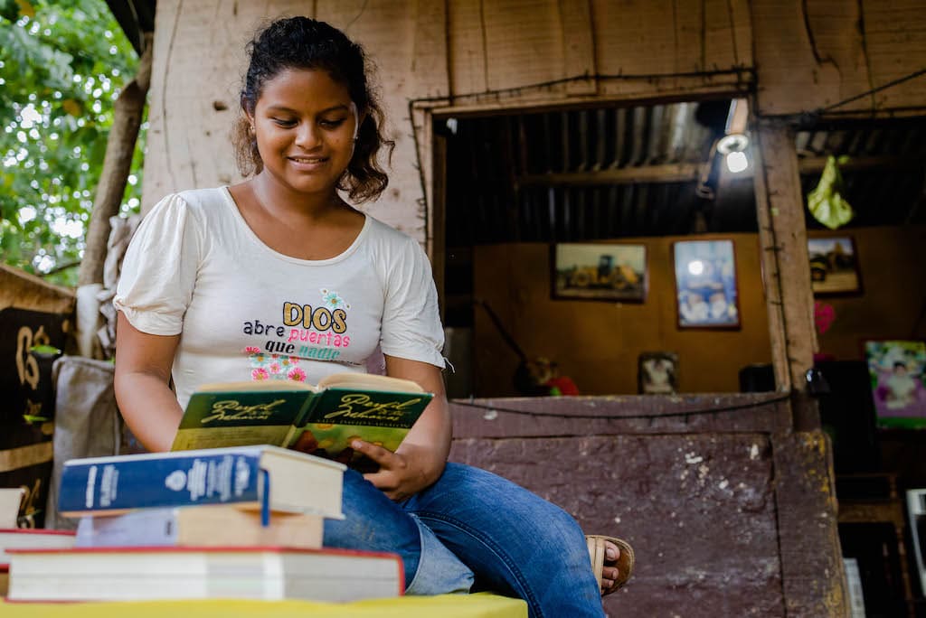 Crisli, wearing a white shirt and blue jeans, is sitting at a table outside her house holding and looking at a book. There are more books stacked in front of her.