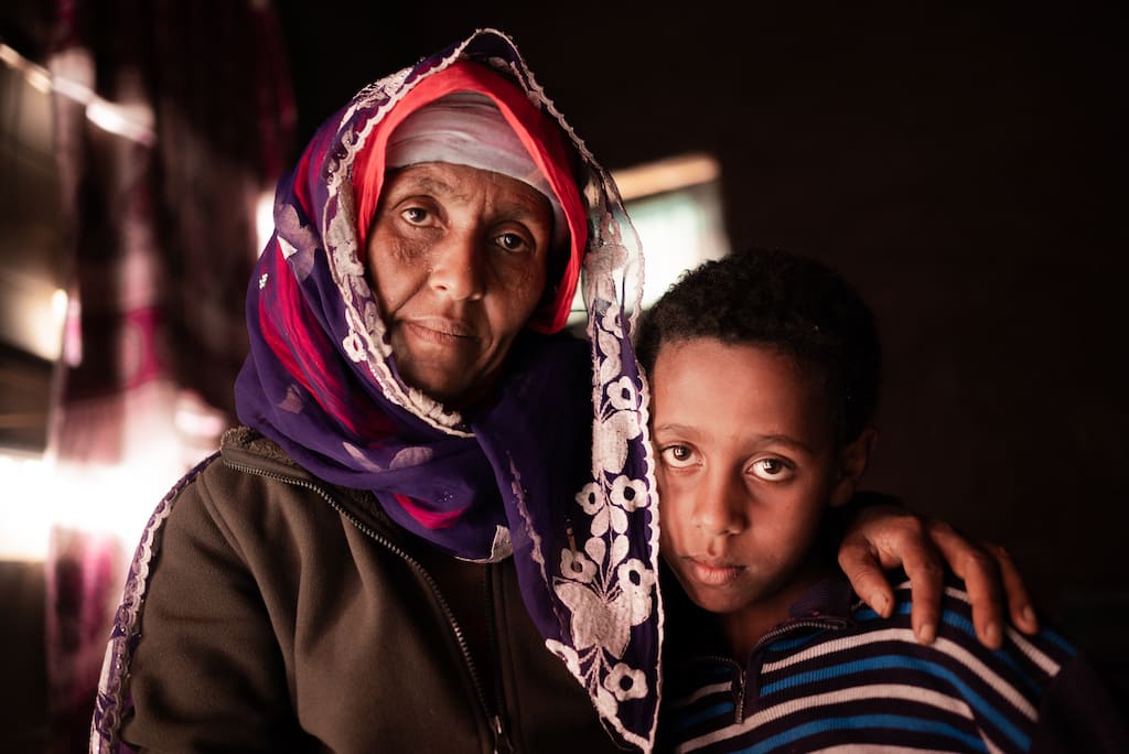 A woman wears a purple and pink scarf on her head while holding her son under her arm beside.