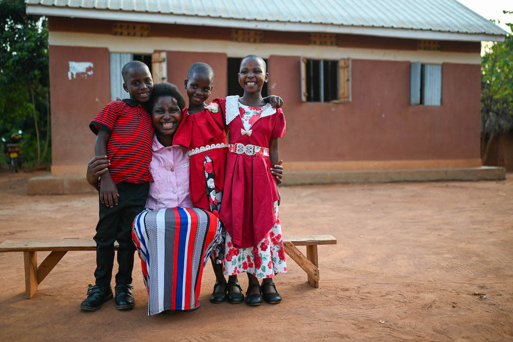 Annett smiles while sitting on a bench with her triplets standing and smiling beside her. They are all wearing red and standing in front of their home.