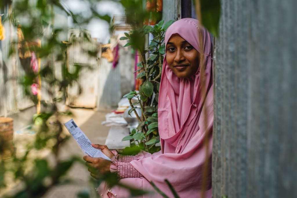 Akhi is wearing a pink dress and head covering. She is sitting in the doorway of her home and is holding a letter from her sponsor.