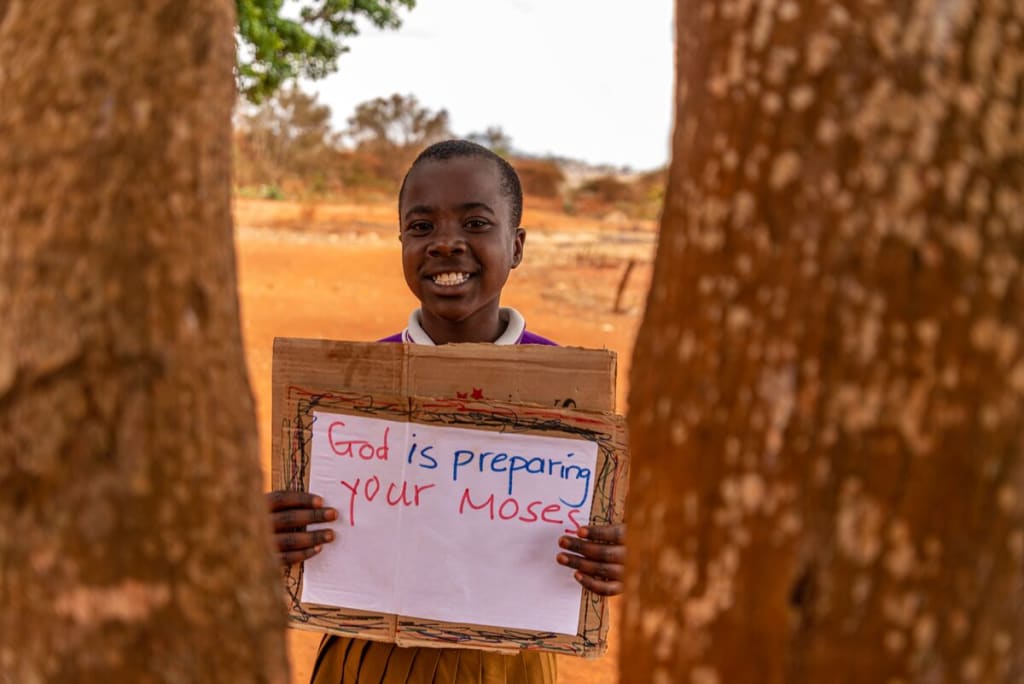 Little boy stands behind two trees holding a sign that says, "God is preparing your Moses."