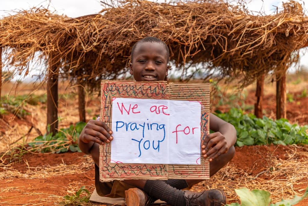Little boy stands under a grass-roof hut holding a sign that says, "We are praying for you!"