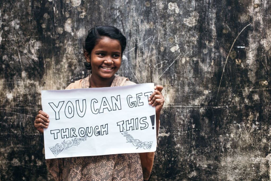 Little girl stands against a cement wall smiling holding a sign that says "You can get through this!"