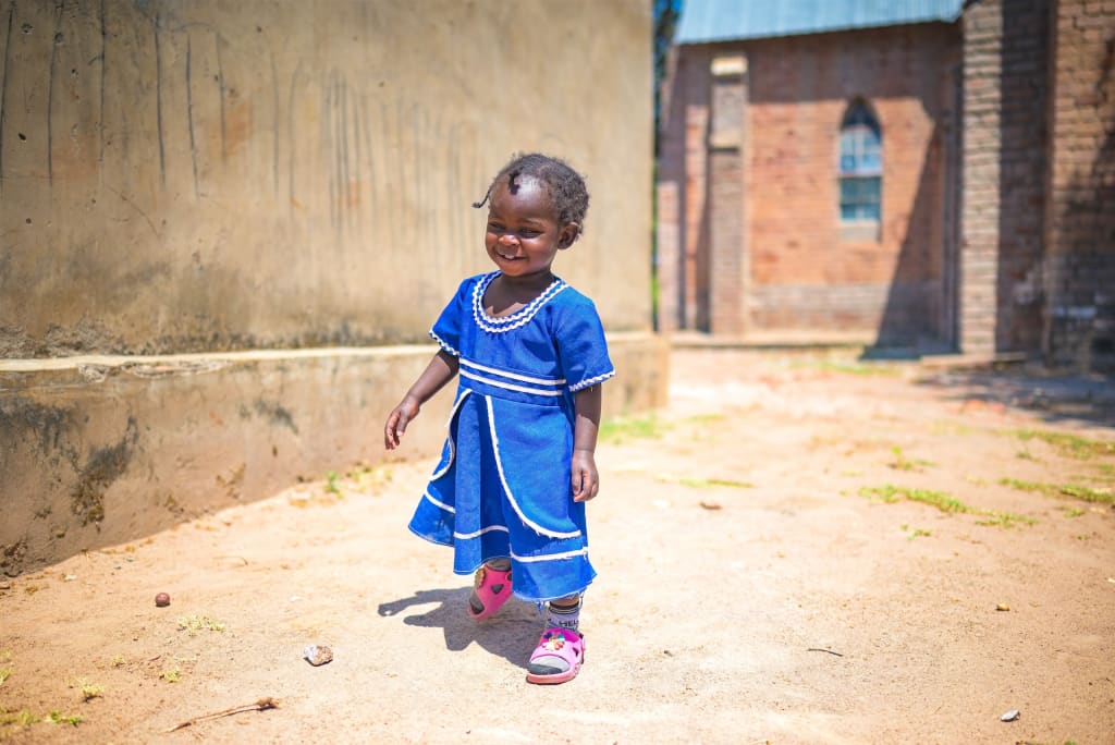 A little girl in a blue dress walks across the sandy ground. She is smiling and wearing red pants