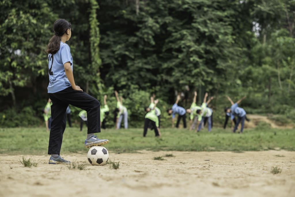 A girl in a blue tshirt and black pants stands looking away with her foot on a soccer ball.