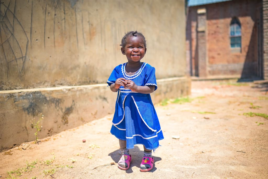 Little girl in a blue dress and pink sandals holds her hands together and gives the camera a big smile.