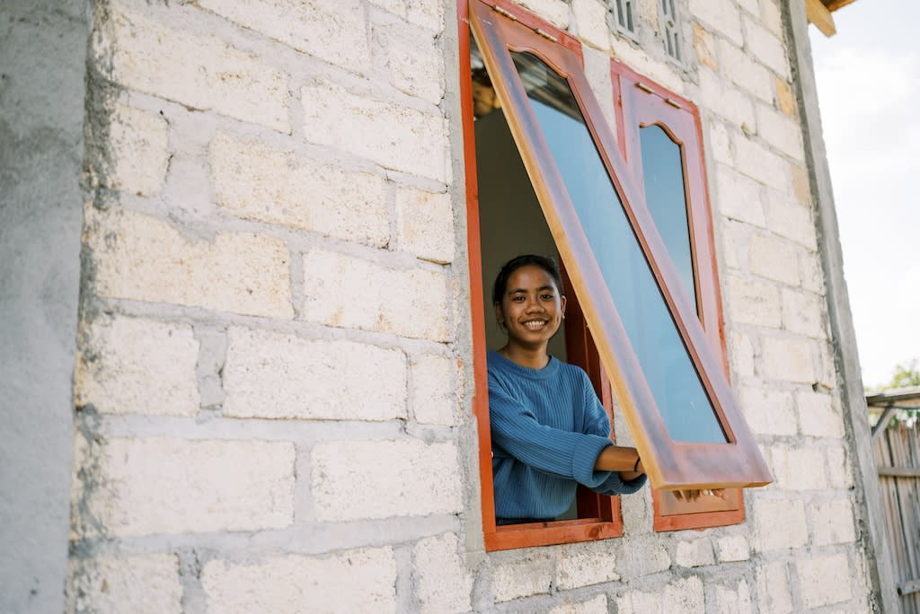 Girl opens a window and smiles at the camera. She is inside a brick building.