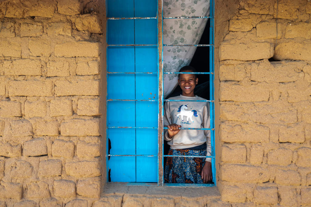 A girl stands inside a home looking out a window with bright blue shutters.
