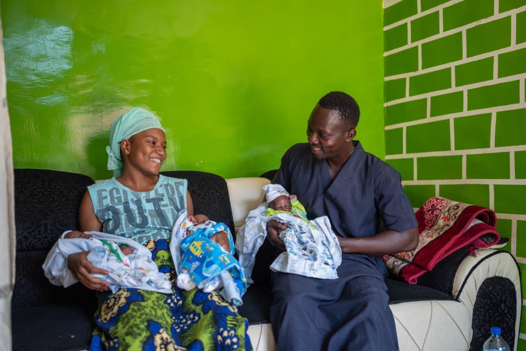 A mother and father hold three newborn babies. They are sitting in their house with a green painted wall.