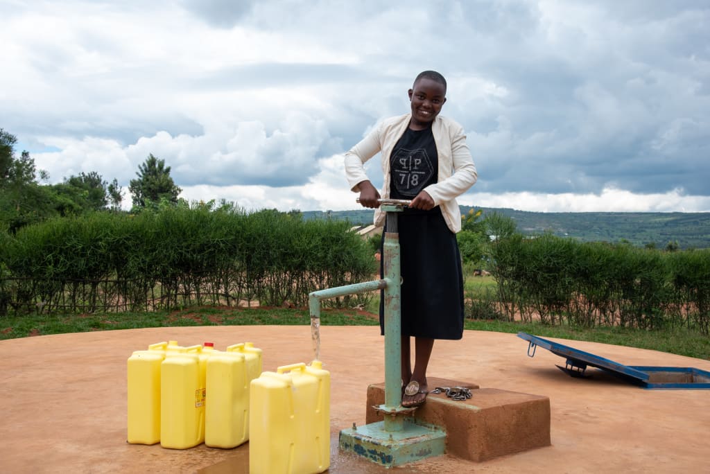 Emeline is wearing a black dress. She is pumping water from the water tank into yellow jerrycans. There are trees in the background.