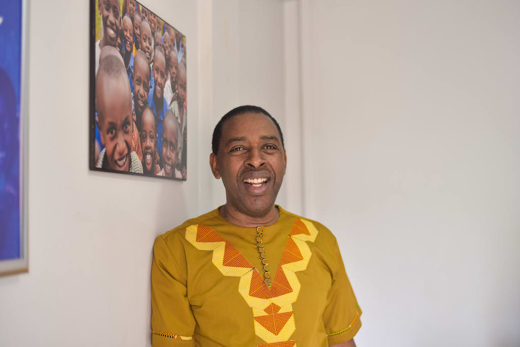 John Nkubana, National Director of Compassion Rwanda, is wearing a gold shirt. He is standing in front of a white wall that has a picture of beneficiary children on it.