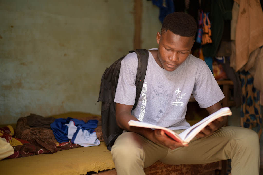 In Burkina Faso, 16-year-old Abdoul is wearing a gray shirt and khaki pants. He is also wearing a black backpack. Abdoul is looking down and reading a book.