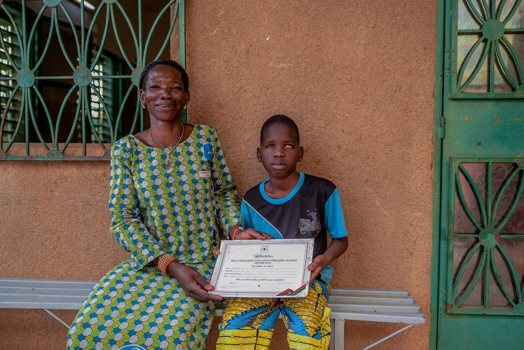 Kader is wearing a blue shirt and yellow patterned pants. He is sitting outside his school next to his mother, Florence, wearing a blue, white, and green dress. They are holding Kader's certificate he received from the president.