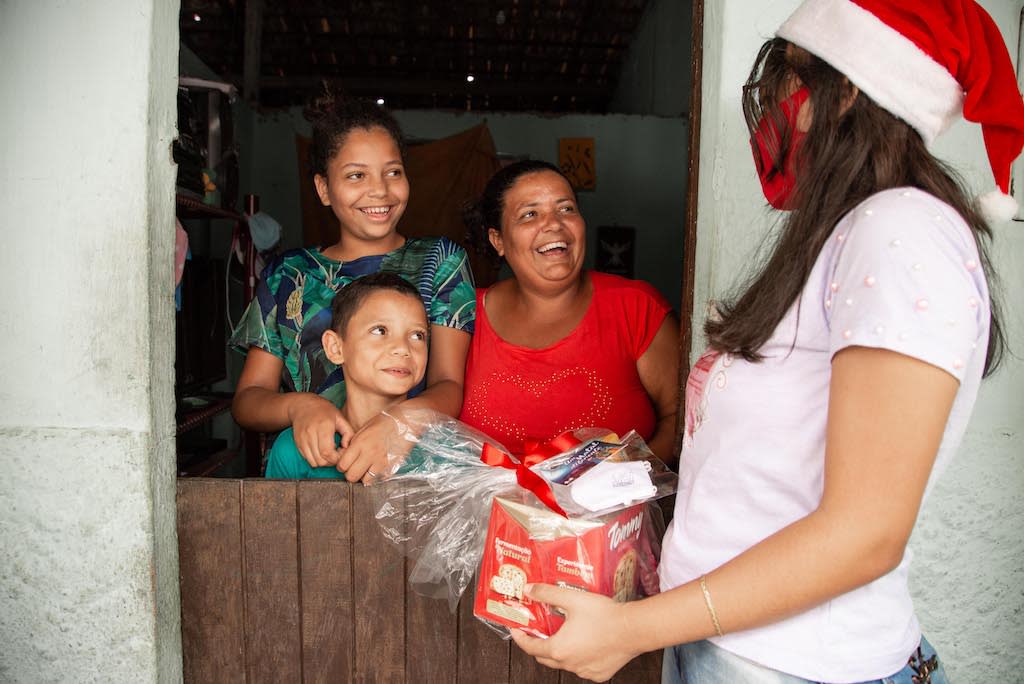 A volunteer wearing a Santa hat and a mask brings gifts to a family of 3 who is smiling behind their door.