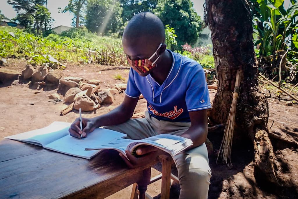 Wearing a cloth protective face mask, Emmanuel sits at a table under a tree, writing in a notebook.