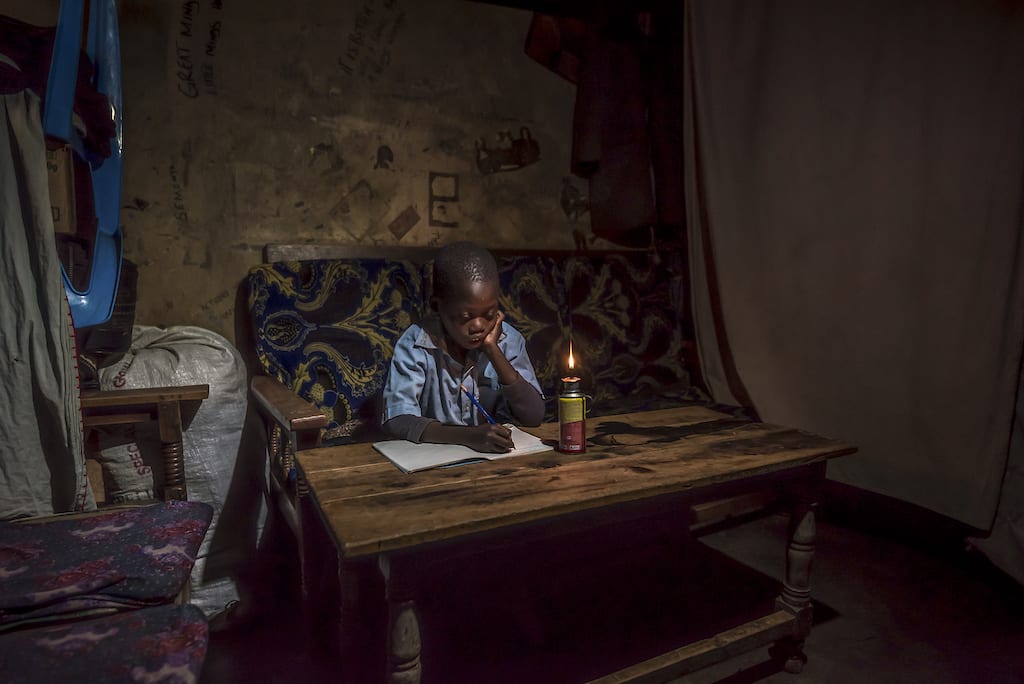 A young boy sits at a wooden table with a notebook in front of him and a kerosene lamp lit nearby.