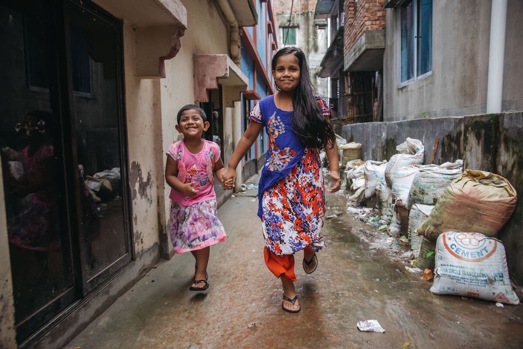 Mim is wearing an orange and purple dress. She is with her sister who is wearing a pink shirt and pink skirt. They are running together down an alley way. There are bags of cement along one side of the alley.
